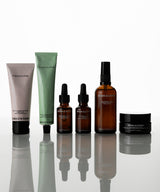 A six-piece skincare set  arranged on a reflective white surface. The set includes a pale pink tube, a mint green tube, two amber dropper bottles, a tall amber spray bottle, and a black jar, all with black caps.