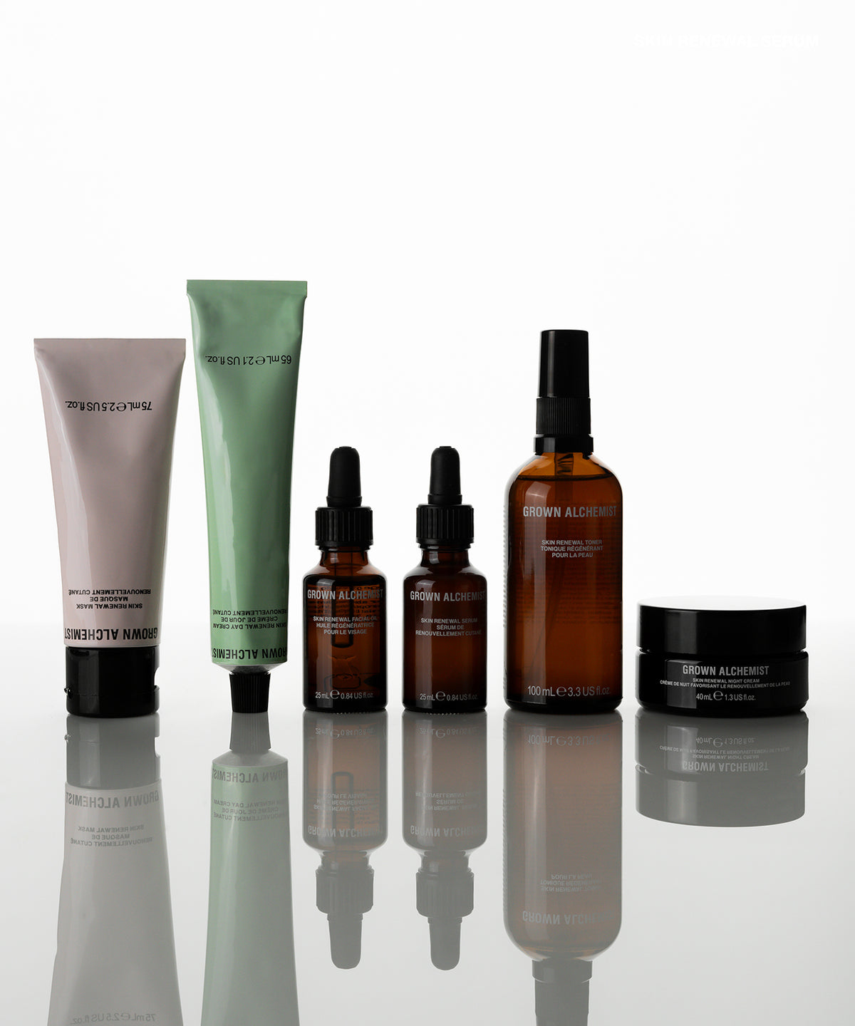A six-piece skincare set  arranged on a reflective white surface. The set includes a pale pink tube, a mint green tube, two amber dropper bottles, a tall amber spray bottle, and a black jar, all with black caps.