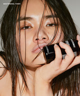 A close-up beauty shot of a woman holding a black cosmetic bottle against her lips. The bottle has white text reading 'SKIN RENEWAL TONER'.