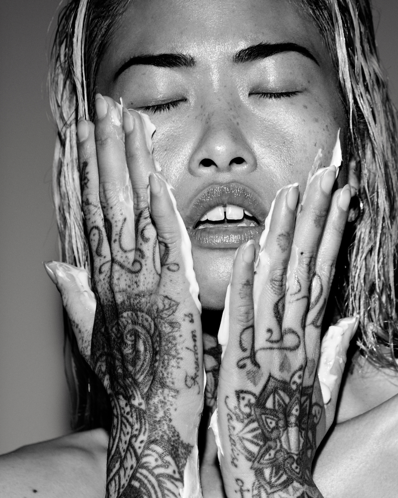 Black and white photo of a person with tattoos on their hands, applying cream to their face