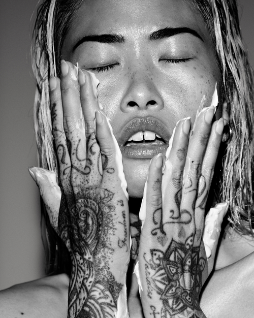 Black and white photo of a person with tattoos on their hands, applying cream to their face